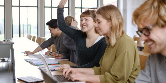 Fünf Studierende sitzen in einem Seminarraum an Tischen. Eine Studentin hebt ihre Hand.